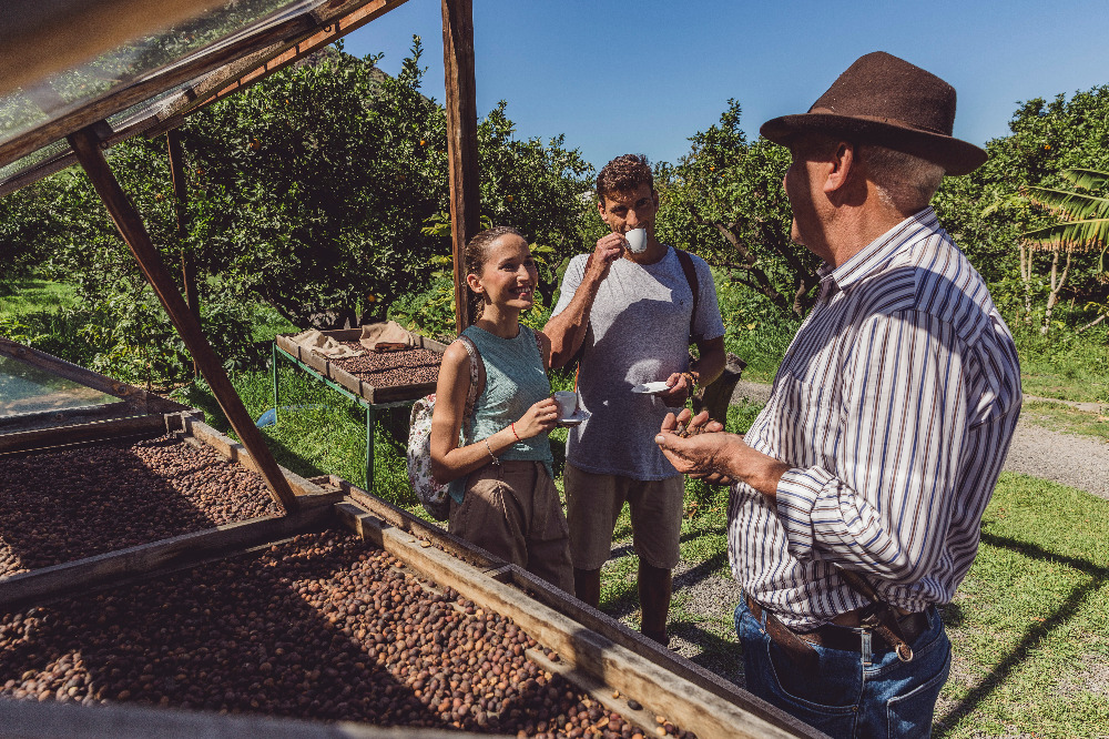 Zwei Besucher:innen verkosten Kaffee und unterhalten sich mit einem Landwirt neben Trockentischen mit Kaffeebohnen.